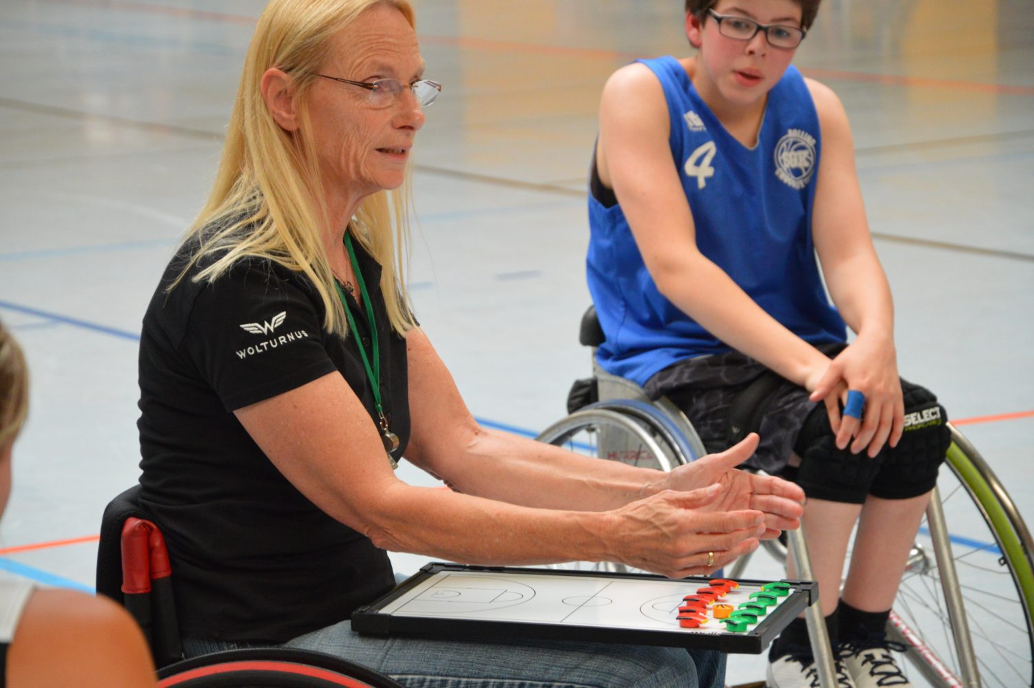 eine Frau im Rollstuhl, ein Taktikbrett mit verschiedenfarbenen Magneten auf dem Schoß