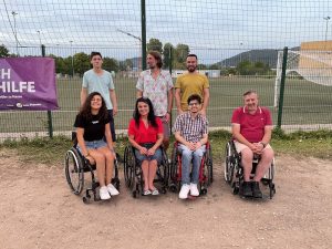 zwei Rollstuhlfahrerinnen, zwei Rollstuhlfahrer und drei Fußgänger stehen auf einem Sportplatz