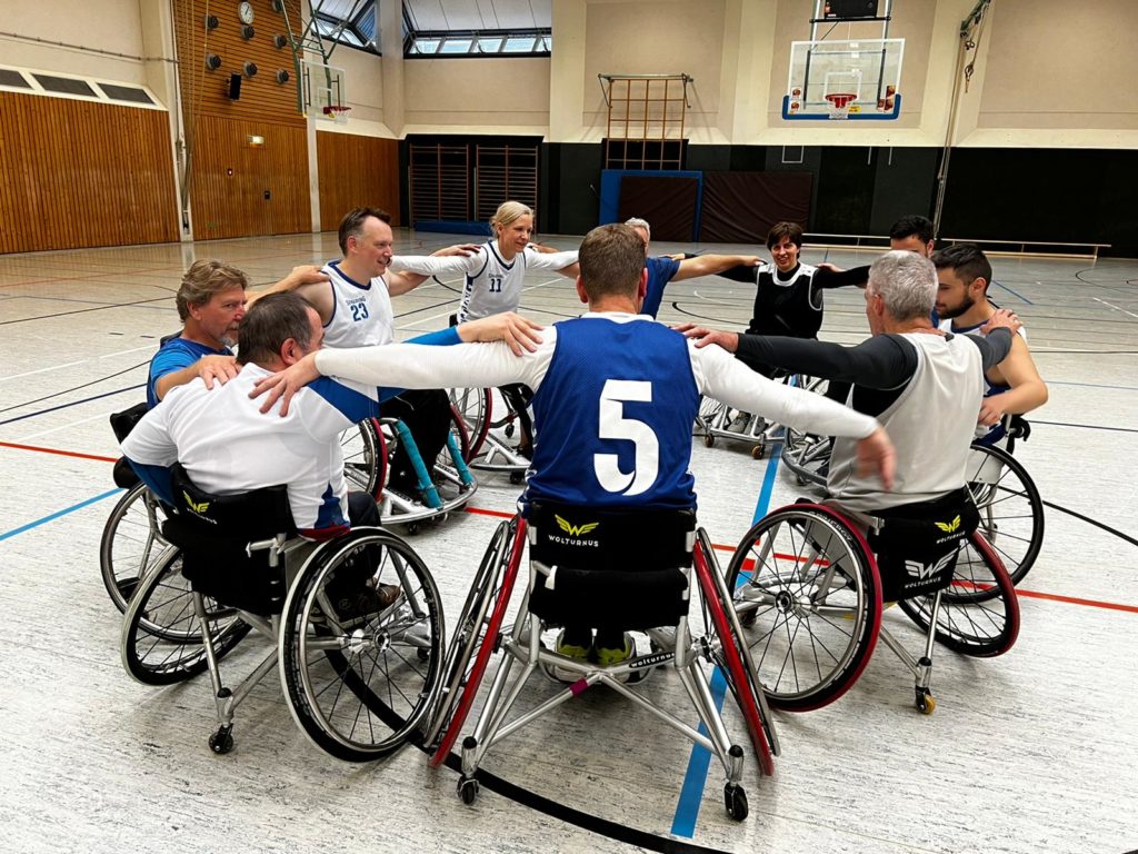 10 Rollstuhlbasketball-Spieler stehen im Kreis. Sie haben die Arme dabei den Mitspielern neben ihnen auf die Schulter gelegt.