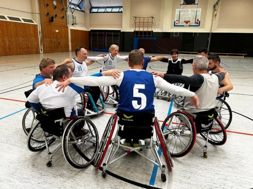10 Rollstuhlbasketball-Spieler*innen stehen im Kreis. Sie haben die Arme dabei den Mitspielern neben ihnen auf die Schulter gelegt.